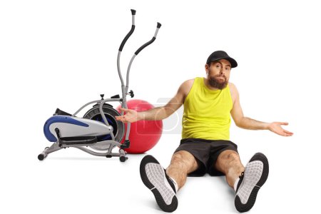 Foto de Hombre barbudo confuso en ropa deportiva sentado en el suelo frente a un equipo de gimnasio aislado en un fondo blanco - Imagen libre de derechos