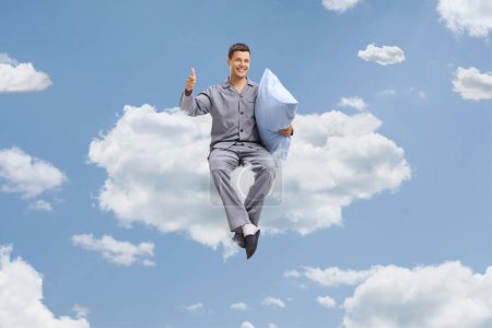 Photo for Young man in pajamas holding a pillow and gesturing thumbs up on a cloud - Royalty Free Image