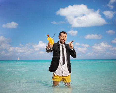 Foto de Hombre de negocios en pantalones cortos de natación con un cóctel y un cigarro de pie en el mar Caribe - Imagen libre de derechos