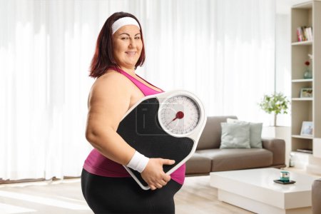 Téléchargez les photos : Femme taille plus en vêtements de sport tenant une balance de poids dans un salon à la maison - en image libre de droit