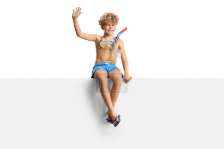 Foto de Niño en pantalones cortos de natación y una máscara de buceo sentado en un panel en blanco y saludando aislado sobre fondo blanco - Imagen libre de derechos