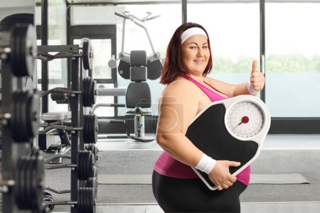 Téléchargez les photos : Femme de taille plus en vêtements de sport tenant une balance de poids dans une salle de gym et les pouces geste vers le haut - en image libre de droit