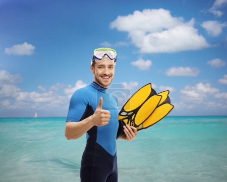 Photo for Man in a snorkeling suit holding swimming fins and showing thumbs up in to the sea - Royalty Free Image