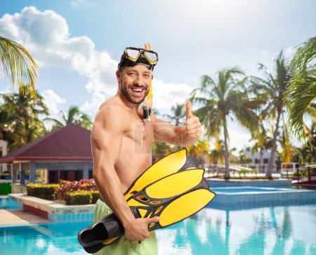 Photo for Cheerful young man in swimwear with a diving mask and snorkeling fins gesturing thumbs up by a swimming pool - Royalty Free Image