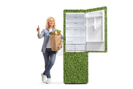 Téléchargez les photos : Femme avec un sac d'épicerie appuyé sur un réfrigérateur vert respectueux de l'environnement et geste pouces vers le haut isolé sur fond blanc - en image libre de droit