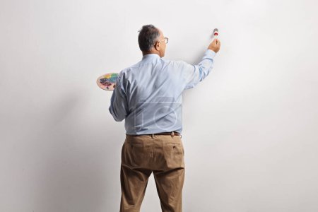 Téléchargez les photos : Vue arrière d'un homme mature tenant un pinceau et une palette devant un mur - en image libre de droit