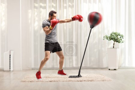 Téléchargez les photos : Plan complet d'un jeune homme fort frappant un stand avec des gants de boxe dans une pièce - en image libre de droit