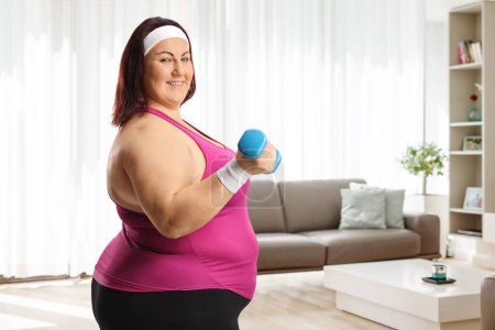 Téléchargez les photos : Femme corpulente tenant un haltère à la maison et souriant à une salle de gym - en image libre de droit
