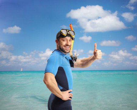 Téléchargez les photos : Jeune homme en combinaison de plongée et masque gestuelle pouces vers le haut dans la mer bleue - en image libre de droit