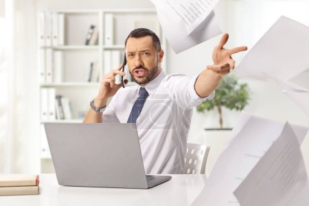 Photo for Angry boss at a desk throwing papers and making a phone call in the office - Royalty Free Image
