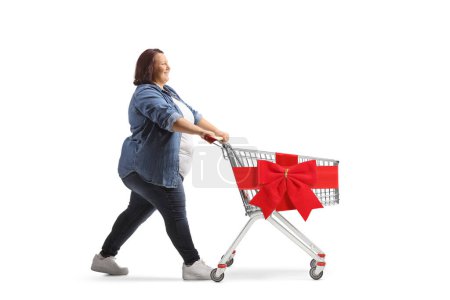 Téléchargez les photos : Photo de profil pleine longueur d'une femme de taille plus poussant un chariot vide avec ruban rouge isolé sur fond blanc - en image libre de droit