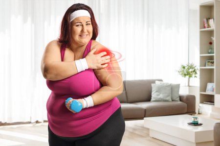 Foto de Mujer de talla grande haciendo ejercicio en casa y sosteniendo su doloroso hombro rojo inflamado - Imagen libre de derechos