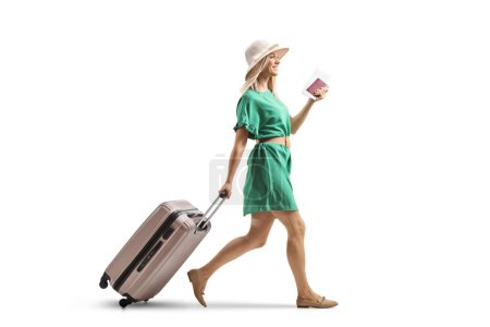 Téléchargez les photos : Jeune femme en robe verte tirant une valise et tenant un passeport isolé sur fond blanc - en image libre de droit