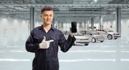 Téléchargez les photos : Mécanicien dans un garage automobile tenant un téléphone portable et pointant du doigt - en image libre de droit