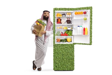 Photo for Saudi arab man in ethnic clothes standing next to an eco-friendly green fridge isolated on white background - Royalty Free Image