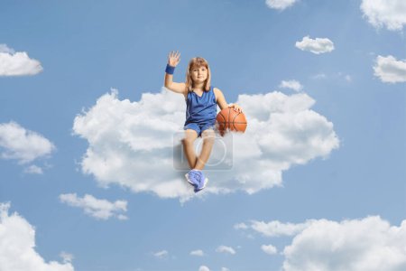 Foto de Niña con una pelota de baloncesto sentada en una nube en el cielo y saludando - Imagen libre de derechos