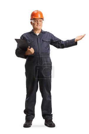 Photo for Male factory worker in an overall uniform and orange helmet holding a clipboard and pointing isolated on white background - Royalty Free Image