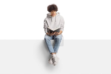 Foto de Joven casual sentado en un panel de blan y leyendo un libro aislado sobre fondo blanco - Imagen libre de derechos