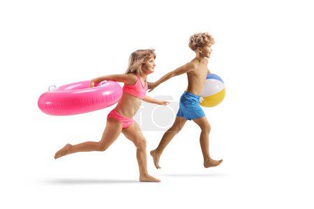 Foto de Niño y niña en traje de baño corriendo y llevando un anillo de baño de goma rosa y una pelota de playa aislada sobre fondo blanco - Imagen libre de derechos