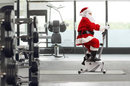 Photo for Santa Claus riding a stationary bicycle at a gym - Royalty Free Image