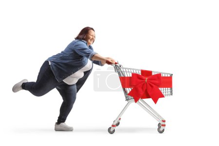 Foto de Foto de perfil completo de una mujer corpulenta corriendo con un carrito de compras vacío con lazo de cinta roja aislado sobre fondo blanco - Imagen libre de derechos