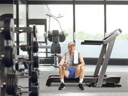 Photo for Elderly man sitting on a treadmill at a gym with a bottle of water and smiling at camera - Royalty Free Image