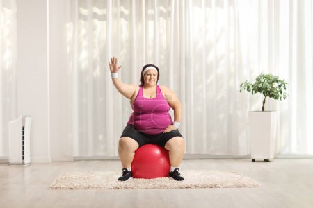 Photo for Corpulent woman sitting on a fitness ball and waving in a room - Royalty Free Image