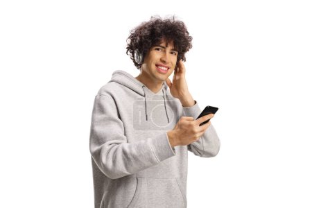 Téléchargez les photos : Guy aux cheveux bouclés avec un smartphone et un casque souriant et regardant la caméra isolée sur fond blanc - en image libre de droit