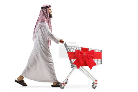 Photo for Full length profile shot of a saudi arab man pushing an empty shopping cart with red ribbon bow isolated on white background - Royalty Free Image