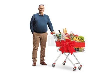 Photo for Full length portrait of a mature man with a shopping cart full of food and tied with a red ribbon isolated on white background - Royalty Free Image