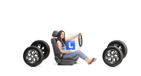Téléchargez les photos : Jeune femme tenant une plaque d'apprenant et un volant assis sur un siège auto et quatre pneus isolés sur fond blanc - en image libre de droit
