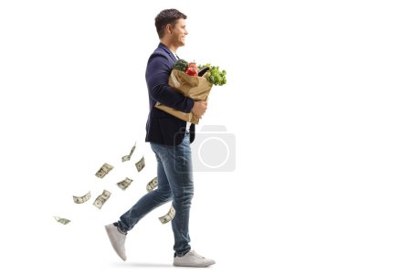 Téléchargez les photos : Plan de profil complet d'un jeune homme marchant et portant un sac d'épicerie avec de l'argent tombant isolé sur fond blanc - en image libre de droit