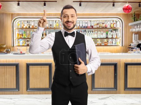 Photo for Cheerful waiter with a bow tie holding a menu and pointing up at a bar - Royalty Free Image