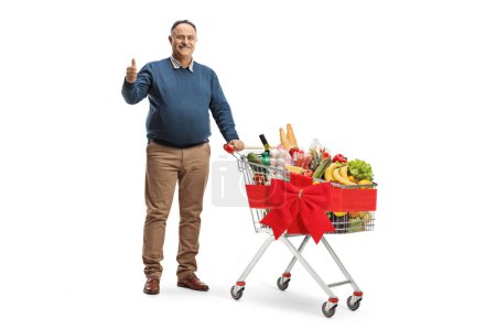 Photo for Full length portrait of a mature man with a shopping cart full of food gesturing thumbs up isolated on white background - Royalty Free Image