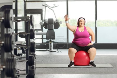 Photo for Corpulent woman sitting on a fitness ball at a gym and waving at camera - Royalty Free Image