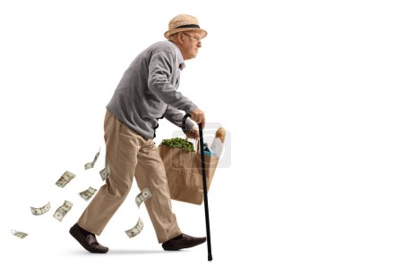 Photo for Full length profile shot of an older man with a cane walking with a grocery bag and losing money isolated on white background - Royalty Free Image