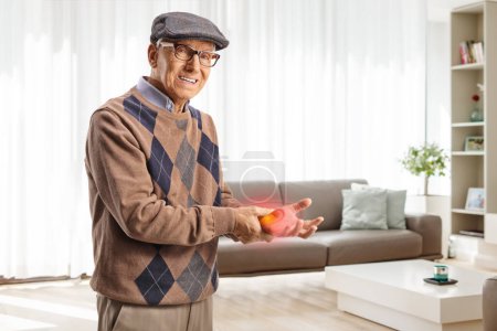 Photo for Elderly man in pain holding his red hand wrist at home in a living room - Royalty Free Image
