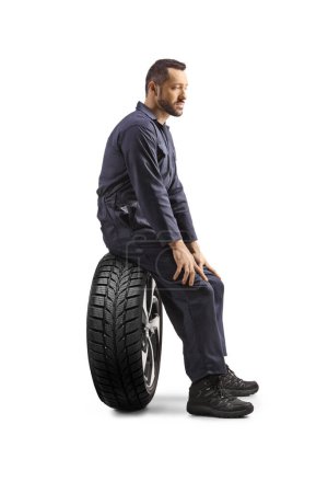 Téléchargez les photos : Vue de profil d'un mécanicien assis sur un pneu isolé sur fond blanc - en image libre de droit