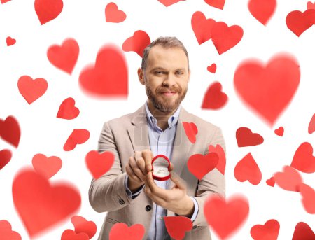 Photo for Man standing under falling hearts and holding an engagement ring in a box isolated on white background - Royalty Free Image