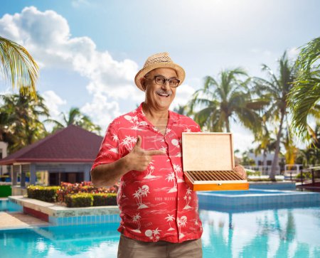 Téléchargez les photos : Touriste mature tenant une boîte de cigares près d'une piscine et pointant - en image libre de droit