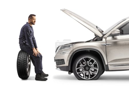 Téléchargez les photos : Vue de profil d'un mécanicien assis sur un pneu de voiture et regardant un VUS avec capot ouvert isolé sur fond blanc - en image libre de droit