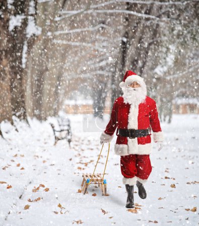 Téléchargez les photos : Père Noël tirant un traîneau en bois dans un parc par une journée enneigée - en image libre de droit