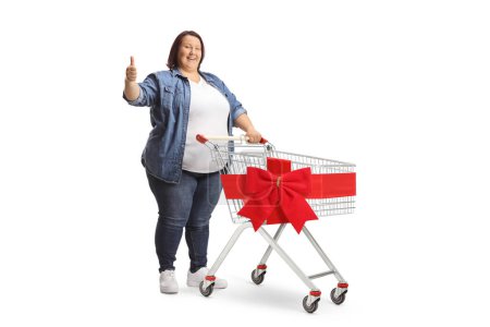Photo for Full length shot of a cheerful corpulent woman standing with a shopping cart and showing thumbs up isolated on white background - Royalty Free Image
