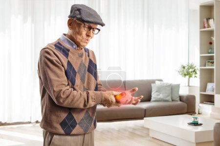 Téléchargez les photos : Homme âgé dans la douleur tenant son poignet enflammé rouge à la maison dans un salon - en image libre de droit