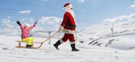 Téléchargez les photos : Photo de profil pleine longueur de Père Noël tirant une petite fille heureuse avec un traîneau en bois sur une montagne enneigée - en image libre de droit