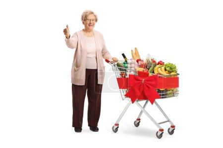 Téléchargez les photos : Femme âgée posant avec un panier plein de nourriture et montrant pouces isolés sur fond blanc - en image libre de droit