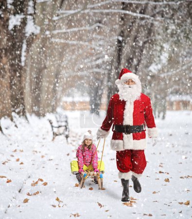 Téléchargez les photos : Père Noël tirant un enfant sur un traîneau en bois dans un parc sur une da enneigée - en image libre de droit