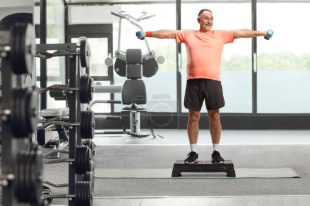 Photo for Full length portrait of a mature man exercising step aerobic and holding dumbbells at the gym - Royalty Free Image