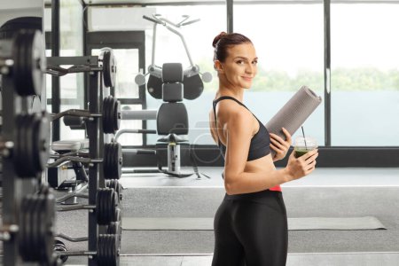 Téléchargez les photos : S'adapter jeune femme tenant un smoothie vert et un tapis d'exercice et se tournant vers la caméra dans une salle de gym - en image libre de droit
