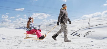 Téléchargez les photos : Homme en vêtements d'hiver tirant une fille avec un traîneau sur une colline de montagne - en image libre de droit